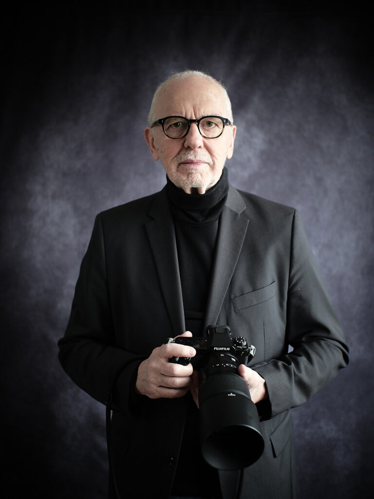 Der Theaterfotograf Peter Levin Schuster steht mit seiner Kamera, einer Fujifilm GFX 100_II vor einer schwarz-weißen Fotowand. Er hat einen schwarzen Anzug und einen schwarzen Rollkragenpullover an.